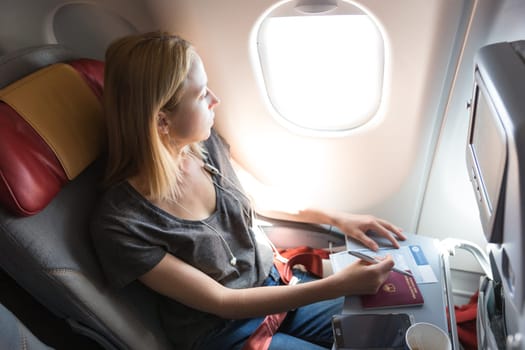 Relaxed casual woman flying on commercial passengers airplane, filling in immigration form, drinking coffee, listening to music.