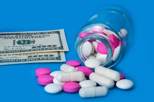 On a blue background, red and white pills scattered from a jar and two bills of one hundred dollars.