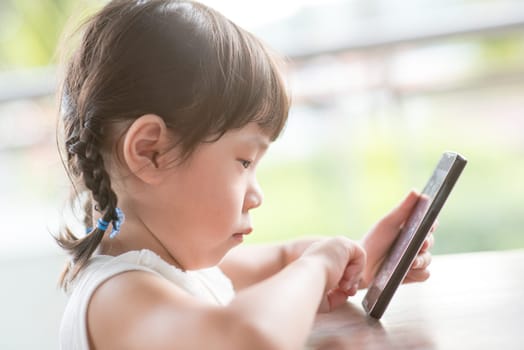Little Asian girl addicted to smart phone at cafe. Natural light outdoor lifestyle.