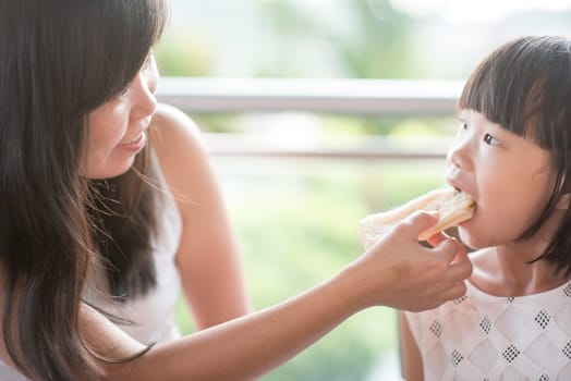 Mom feed bread to her child at cafe. Asian family outdoor lifestyle with natural light.