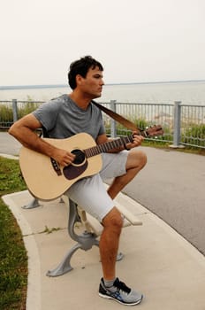 A lonely young man sitting on a bench on the lake and playing his .guitar, looking away over the lake.