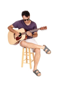 A young man sitting in jeans and sunglasses, playing his guitar, looking.down, isolated for white background.