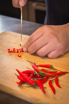 Hand slicing Chilli pepper with Knife on chopping board on wooden background.