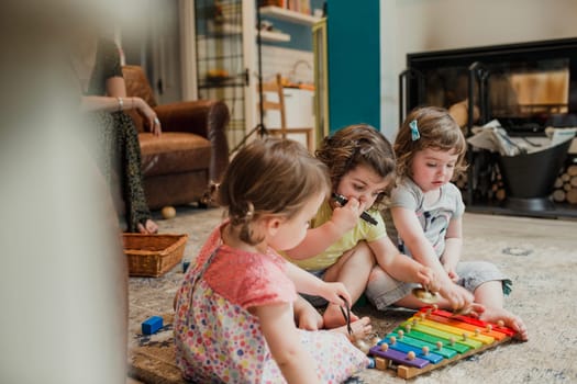 Side view of three little girls sitting on the floor in a living room. The little girls are playing with instruments and enjoying themselves. An unrecognisable person is sat behind them on the sofa watching and keeping an eye on them.