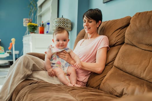 Mid adult mother sat on a living room sofa. The baby is sat up on the mothers lap being winded after having a bottle. The baby is looking at the camera and the mother is smiling at the baby.