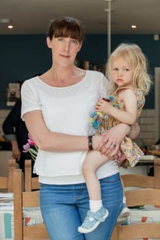 Portrait of a mid adult mother and her daughter looking at the camera. The mother is holding her daughter at the side of her. They are standing indoors in a kitchen.