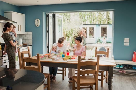 Wide angle view of three mid adult mothers sitting at a kitchen table talking. Two of the mothers have their children with them and enjoying some snacks.