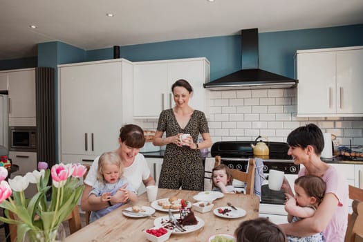Three mid adult women in a kitchen with their daughters. Two of the women are sitting down with their daughters sitting on their laps while they eat their lunch. One mid adult woman is standing with a cup of tea and smiling.