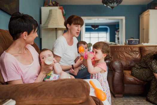 Side view of two mothers sitting on a a sofa in a living room. One mid adult woman is feeding her baby with a bottle of milk an her other daughter is stood in front of her with a sippy cup of juice having a drink.