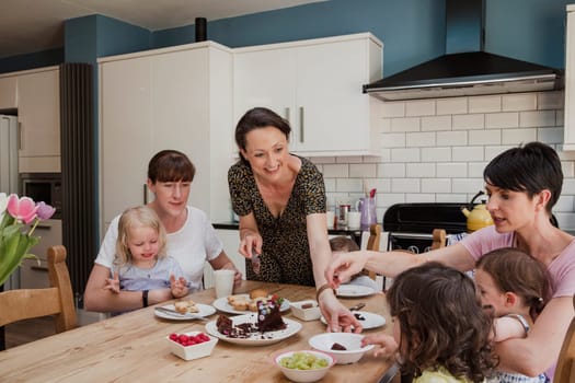 MId adult woman handing a slice of cake to a little girl sat at the dining table. There are two other mid adult women sat at the table with their children enjoying the sweet treats.
