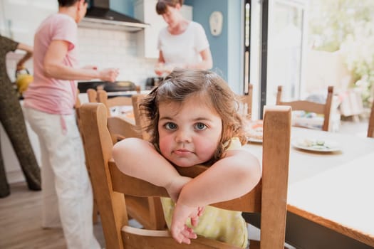 Little girl sitting hanging off the back of a dining table chair in the kitchen. The little girl is sat waiting for her lunch to be made.