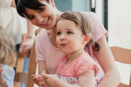 Side view of a little girl sitting on her mothers lap. She is trying a new food and pulling a face while trying it. Her mother is smiling and laughing while looking at her.