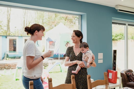 Two mid adult women standing in the kicthen at home and socialising. One woman is holding her baby son. They are talking and laughing together.