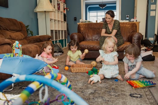 Small group of little girls playing with toys in the living room. One mid adult woman is stiiting watching and keeping an eye on them as they play together.