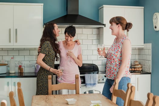 Three mid adult women standing in a kicthen enjoying a glass of wine. The women are all standing around the stove in the kitchen, talking and laughing.