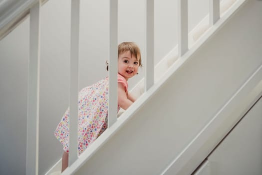 Side view of a staircase in a house. There is a toddler girl climbing up the stairs. She is turning her head and smiling while looking at the camera.