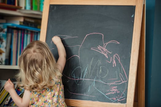 Rear view of a little girl drawing and writing on a blackboard. The little girl is reaching up and drawing, using different coloured chalk on the black board.