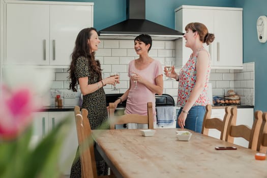 Three mid adult women standing in a kicthen enjoying a glass of wine. The women are all standing around the stove in the kitchen, talking and laughing.