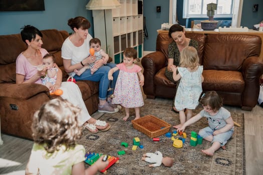 Wide angle view of a group of children and their mothers in a living room. The children are playing with musical instruments and toys. One mother is sat feeding her child a bottle of milk.