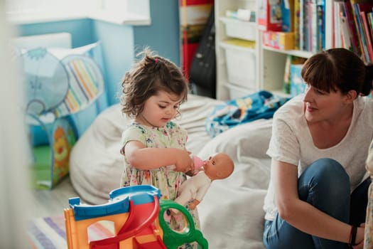 Little gilr standing in a room with her mother and using a toy bottle of milk to feed her toy baby doll. The little girls mother is sitting down watching her play with her dolls.
