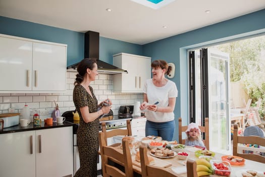 Side view of two mid adult friends standing in a kitchen making lunch for the home day care. They are talking and laughing with eachother, one little girl is sitting on a chair waiting for her lunch.