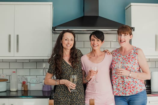 Three mid adult women standing in a kicthen enjoying a glass of wine. The women are all standing around the stove in the kitchen, looking at the camera and smiling.