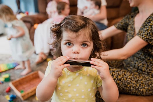 Front view of a little toddler girl looking at the camera. She is having fun and playing the harmonica. In the background ther are other children playing and her mother is sat down next to her.