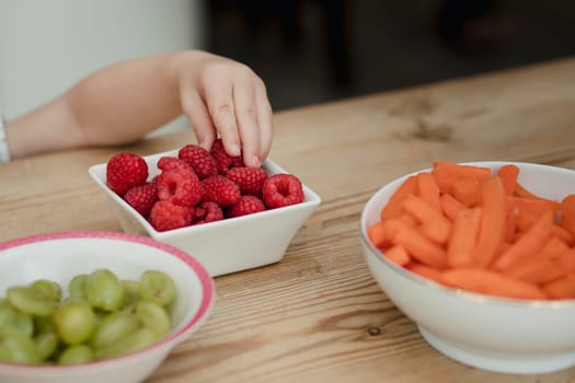 Part of a little girls arm reaching over a kitchen counter to grab some raspberries from a bowl with some other snacks in some bowls close.