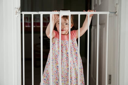 Front view of a little girl standing on the other side of a baby saftey gate. The little girl is holding onto the top of the saftey gate and looking at the camera over the top.