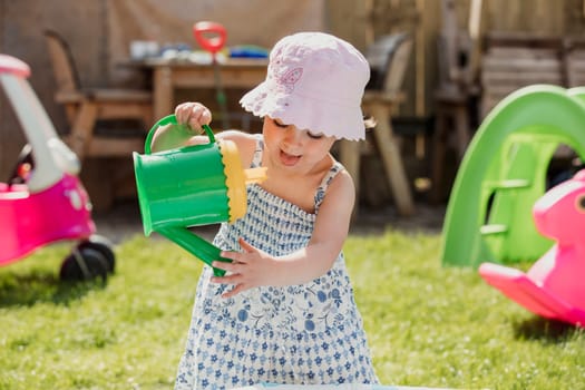 Front view of a little girl standing in the back garden with casual clothing and a sun hat on. She is playing with a toy watering can and tipping it to make the water come out.