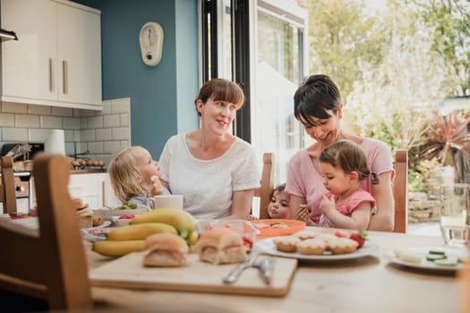 Two mid adult women sitting in the kitchen at the dining table with their two daughters on their laps. The two little girls are enjoying their lunch while their mothers talk and smile.