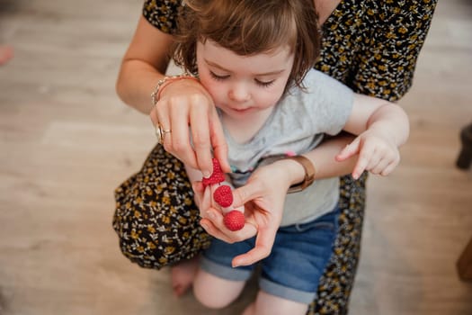 Front view of a little girl playing with some raspberries and putting them onto her fingers. Her mother is kneeling behind her helping and laughing with her.