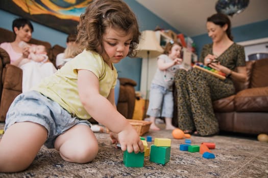 Low angle view of a little girl sitting in the living room with her mother and her mothers friends. The little girl is sitting on the floor playing with some building blocks.
