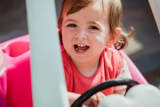 Little girl looking up at the camera and smiling with her mouth open. She is sat in a toy car, excited to play and have fun.