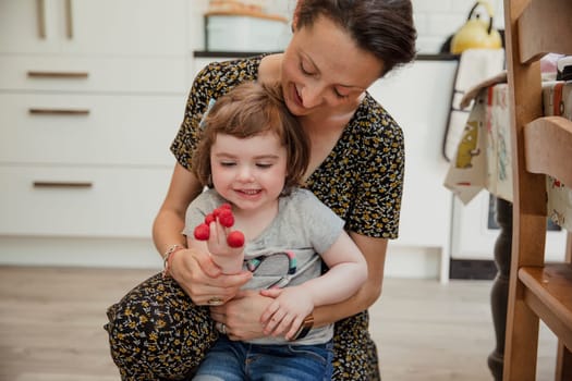 Front view of a little girl playing with some raspberries and putting them onto her fingers. Her mother is kneeling behind her helping and laughing with her.