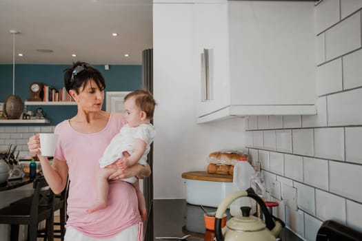 Front view of a mother holding her baby daughter while standing in the kitchen. The woman is holding a cup of tea while carrying her daughter on her side.