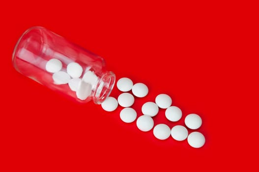 On a red background, white tablets are scattered from a glass bottle. View from above.