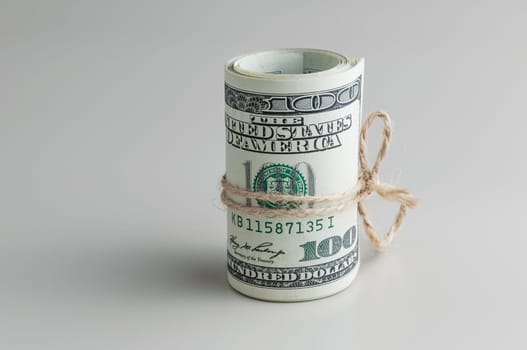 A roll of one hundred dollar bills tied with a rope on a gray background. Vertically. Side view.