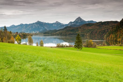 Landscape view with green meadow in the foreground, the Weissensee in the middle area and the first alpine peaks with the Säuling as the highest elevation in the background in the evening light mood.