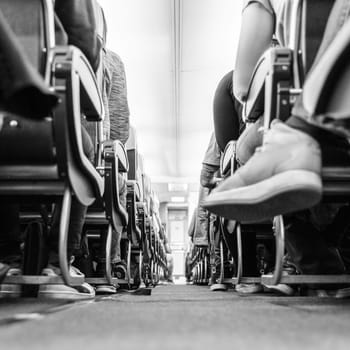 Low agle view of passenegers commercial airplane aisle with passenegers sitting on their seats while flying. Black and white image.