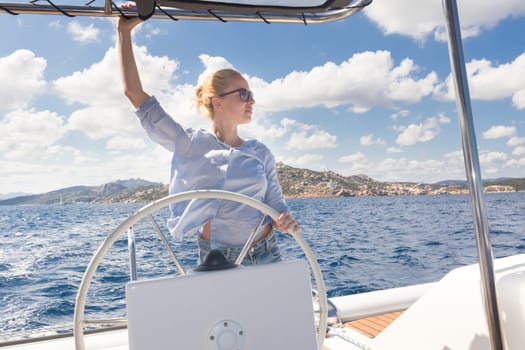 Attractive blond female skipper navigating the fancy catamaran sailboat on sunny summer day on calm blue sea water. Luxury summer adventure, active nautical vacation.