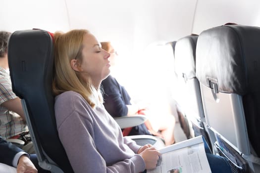 Tired blonde casual caucasian woman napping on seat while traveling by airplane. Commercial transportation by planes.