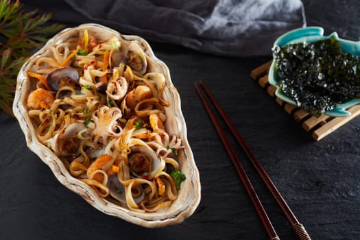 Fried Japanese noodles with a variety of seafood including baby squid, calamari, clams, shrimp and shellfish served in a shell viewed high angle on black slate with copy space