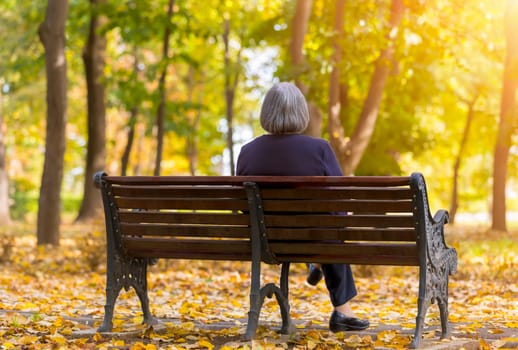 Elderly woman sitting on a bench in autumn park. Lonely aged woman sitting on bench in autumn park lookes at trees.
