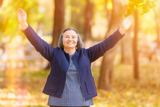 Casual senior woman with arms outstretched standing in autumn park.