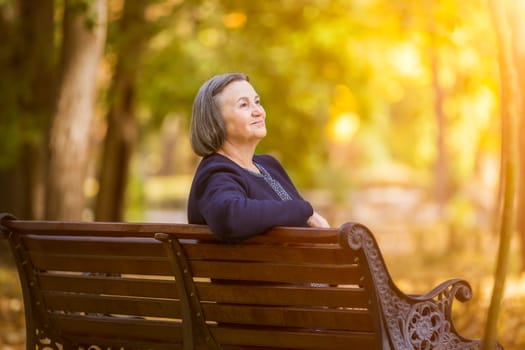 Elderly woman sitting on a bench in autumn park. Aged woman sitting on bench in autumn park looks and smiling at the sun.
