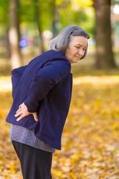 Elderly woman with lower back pain in autumn city park. Unhappy senior woman while feels pain from backache walking in autumn park.