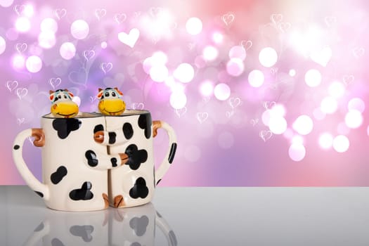 Valentine day concept. Sweet cows hug each other as a form of two mugs on a bright table in front of a love hearts bokeh background. Template for your display product montage.