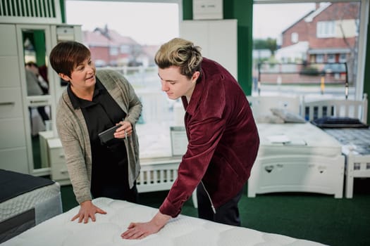 A saleswoman is assisting a young man as he shops for a new matress. She is talking to him while he tests a display.