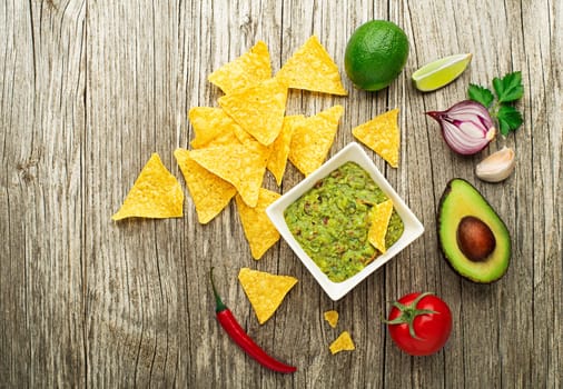 Guacamole sauce with fresh ingredients and corn nachos chips on wooden background